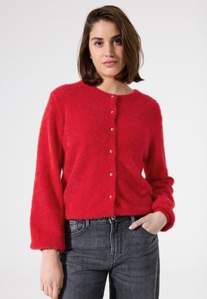 Rode, donzige cardigan gemaakt van zacht materiaal met een ronde hals, zeven knopen aan de voorkant en losse mouwen. Gegdraagd met donkere jeans.