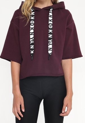 Kvinde iført en kort bordeaux hættetrøje med brede ærmer og sort-hvide snore, kombineret med sorte leggings.