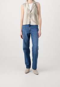 Beige ärmelloses Oberteil mit Kragen und Schleife vorne, kombiniert mit hochtaillierten blauen Jeans und hellen Absatzschuhen.