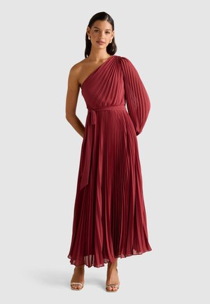 Donna che indossa un maxi abito plissettato bordeaux a spalla scoperta con una lunga manica a palloncino e una cintura annodata in vita, abbinato a tacchi aperti dorati.