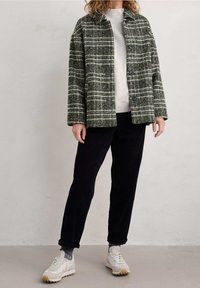 Grün-weiße karierte Jacke mit Kragen, zwei Knöpfen und großen Taschen, getragen über einem hellgrauen Pullover und schwarzen Cordhosen.