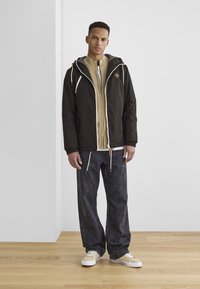 Black hooded jacket with white drawstrings over a beige zip-up sweater. Gray denim pants and light sneakers with a white sole.