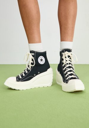 Baskets montantes noires avec une semelle en caoutchouc blanche et un talon compensé. Comprennent des lacets, des œillets et un logo circulaire sur le côté.