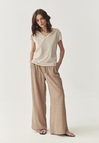 Tricou cu decolteu în V, cu dungi bej și maro, asortat cu pantaloni largi, texturati, bej. Sandale cu bretele subțiri completează ținuta.