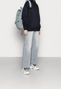 Granatowy hoodie z przednią kieszenią, jasnoszare dżinsy o szerokich nogawkach oraz masywne białe sneakerzy z czarnymi i jasnoniebieskimi akcentami. Szary plecak z paskami.