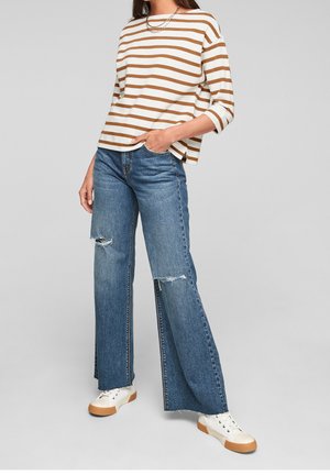 Kvinde iført brun- og hvidstribet langærmet top, slidte brede blå jeans og hvide sneakers med gummisåler.