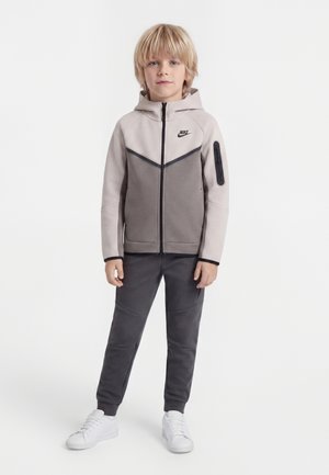 Blond chłopiec stoi, ubrany w dwukolorową szarą bluzę Nike z kapturem i zamkiem, ciemnoszare spodnie oraz białe buty sportowe, na jednolitym białym tle.