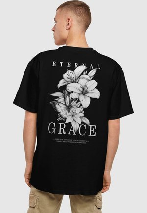 Sort oversized t-shirt med et detaljeret hvidt blomstermotiv på ryggen, med ordet "GRACE" og dekorativ tekst nedenfor.