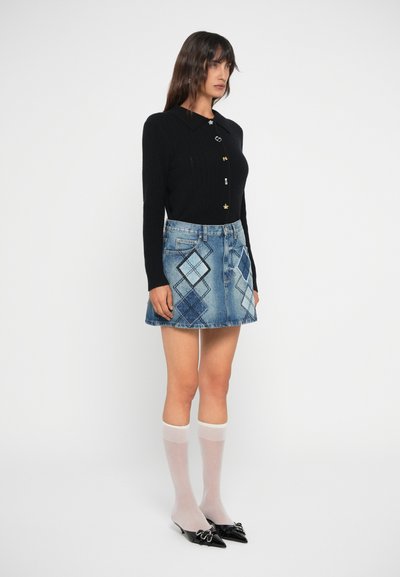 Suéter negro de manga larga con relieve y botones dorados combinado con una falda de mezclilla azul con patrones de diamantes, calcetas blancas hasta la rodilla y zapatos negros de punta.