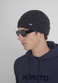 Donkergrijze geribbelde gebreide muts met een zilveren logo-accent, gecombineerd met zwarte rechthoekige zonnebril. Gekocht met een marineblauwe hoodie.