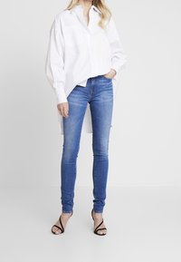 Camisa blanca de gran tamaño con dos bolsillos en el pecho, combinada con unos pantalones vaqueros ajustados de denim azul y tacones negros con tiras.