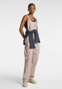 Beige Latzhose mit verstellbaren Trägern, Seitentaschen, einem Bindedetail mit einem blauen gesteppte Stück und hellbeigen Schuhen, vor einem weißen Hintergrund.