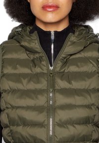 Gilet rembourré vert olive avec une fermeture éclair à l'avant et une capuche. Présente des sections matelassées horizontales et une texture de col noir côtelé.