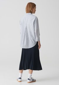 Camicia a righe con orlo arrotondato, abbinata a una gonna blu a pieghe. Le scarpe sono bianche con dettagli blu e suole di gomma, indossate con calzini bianchi.