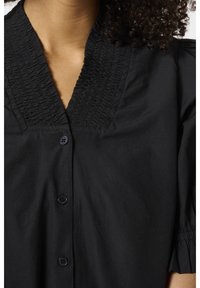 Femme portant une blouse noire à boutons avec un col en V texturé et des manches bouffantes resserrées aux poignets.