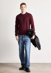 Μπορντό πλεκτό πουλόβερ με στρογγυλή λαιμόκοψη, συνδυασμένο με ανοιχτό μπλε τζιν κανονικής γραμμής. Μαύρο μπουφάν μπομμπέρ κρατημένο στο αριστερό χέρι, μαύρα παπούτσια.