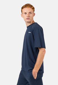 Námorně modré bavlněné tričko s krátkým rukávem, kulatým výstřihem a malým bílým logem na hrudi. Hladká textura, volný střih.