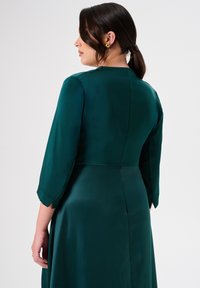 Abito verde smeraldo con maniche a tre quarti, texture in satin liscio, busto aderente e dettagli sulla cucitura dritta sul retro.