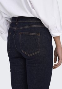 Jeans in denim blu scuro con vestibilità slim, caratterizzati da cuciture arancioni, due tasche posteriori e un'etichetta in pelle del brand sulla vita.