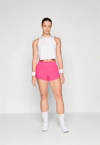 Top atlético branco cortado com decote redondo, combinado com calções rosa de cintura alta. Braceletes de pulso brancas e sapatilhas completam o conjunto.