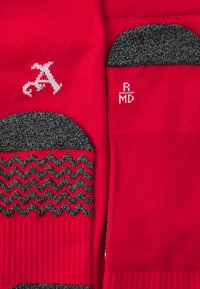 Chaussettes tricotées rouges avec un motif en zigzag gris, présentant un logo blanc et du texte. La texture est lisse, avec des accents côtelés en haut.