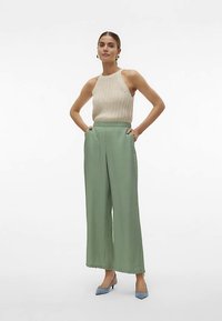 Πλεκτό μπεζ τοπ halter σε συνδυασμό με ψηλόμεσα, φαρδιά παντελόνια σε μέντα πράσινο χρώμα. Το μοντέλο φοράει ανοιχτό μπλε μυτερά τακούνια. Απλός σχεδιασμός, λεία υφή.