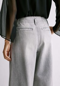 Kobieta w jasnoszarych, szerokich spodniach z wysokim stanem i przezroczystej czarnej bluzce z długim rękawem, pokazana od tyłu na wysokości talii.