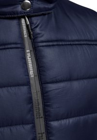 Veste rembourrée bleu marine avec une finition texturée, présentant une fermeture éclair grise et un détail de logo. Le design comprend des coutures horizontales et un col haut.