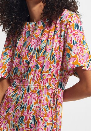 Kobieta ubrana w sukienkę z krótkim rękawem, taliowaną, z pomarańczowym tłem i jasnoróżowym wzorem kwiatowym.