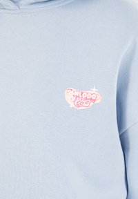 Sudadera azul claro de tejido suave, con un logotipo impreso en rosa en el pecho. El diseño incluye formas redondeadas y acentos.
