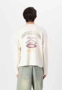 Top de algodón de manga larga en color crema con escote redondeado y hombros caídos. Presenta un estampado gráfico en la parte posterior en texto metálico y en negrita.