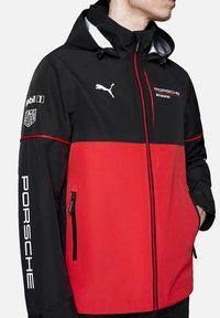 Czarna i czerwona kurtka z zapięciem na zamek z przodu, kapturem i bocznymi kieszeniami. Ozdobiona białymi detalami marki Porsche oraz Puma. Gładki, lekki materiał.