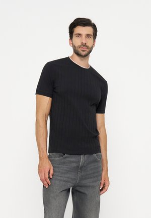 Homme portant un t-shirt noir côtelé à manches courtes et un jean gris, debout devant un fond blanc uni.