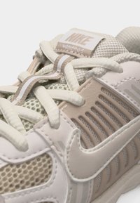 Κοντινή λήψη παπουτσιού Nike Vomero 5 σε μπεζ και λευκό, που δείχνει τα κορδόνια, το δίχτυ και το λογότυπο στο πλευρό σε υφασμάτινη επιφάνεια με υφή και συνθετικά υλικά.