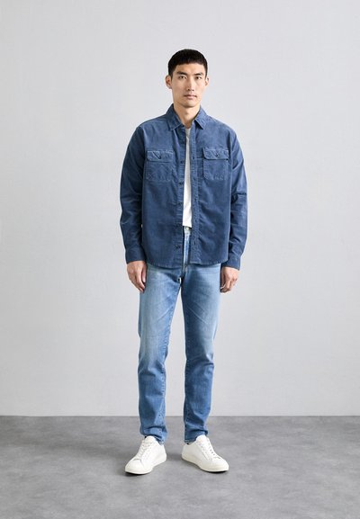 Camisa de color azul oscuro con dos bolsillos en el pecho, combinada con jeans azul claro y zapatillas blancas, sobre un fondo gris neutro.