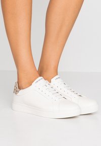 Närbild av ben som bär vita Emporio Armani-sneakers med beige logodetalj på hälen och vita skosnören mot en ljus bakgrund.