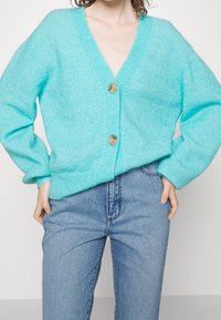 Cardigan en tricot turquoise avec un col en V, fermeture à boutons et manches longues, associé à un jean bleu clair. Texture douce et coupe décontractée.