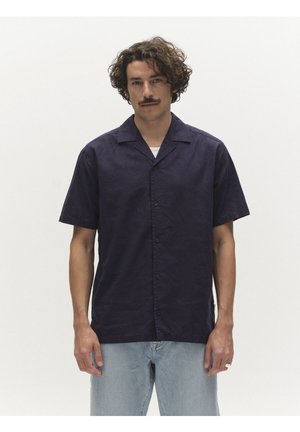 Mies, jolla kiharat hiukset ja viikset, pukeutunut laivastonsiniseen lyhythihaiseen nappipaitaan valkoisen aluspaidan ja vaaleansiniset farkut päällä, seisoo.