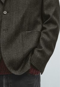 Blazer grigio scuro a spina di pesce con colletto a revers, due bottoni, tasche laterali, indossato sopra un maglione bordeaux lavorato a maglia, tessuto strutturato.