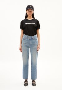 Camiseta negra con el texto "ARMEDANGELS", jeans rectos de color azul claro y gorra negra con detalle de cinta. Zapatos negros con diseño recortado.