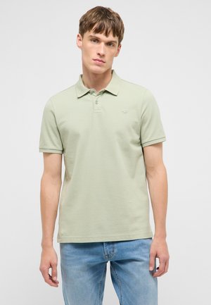 Junger Mann, der ein hellgrünes kurzärmliges Poloshirt und blaue Jeans trägt, blickt mit neutralem Ausdruck nach vorne.