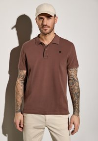 Camisa polo marrón de manga corta con cuello y tapeta de tres botones. Presenta un pequeño logo negro en el pecho. Modelo llevando una gorra clara.
