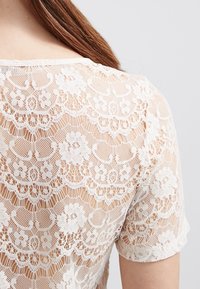 Blusa de encaje con patrón floral, mangas cortas y un diseño semi-translúcido en blanco roto, con un borde ondulado en el dobladillo y el escote.