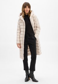 Beige Pufferjacke mit Kapuze, gestepptem Design, Frontreißverschluss und seitlichen Taschen, kombiniert mit schwarzen Wildlederhosen und schwarzen Stiefeletten mit Absatz.