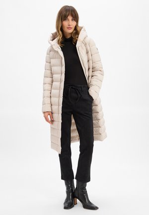 Beige puffer coat with a hood, quilted design, front zip, and side pockets, paired with black suede pants and black heeled boots.