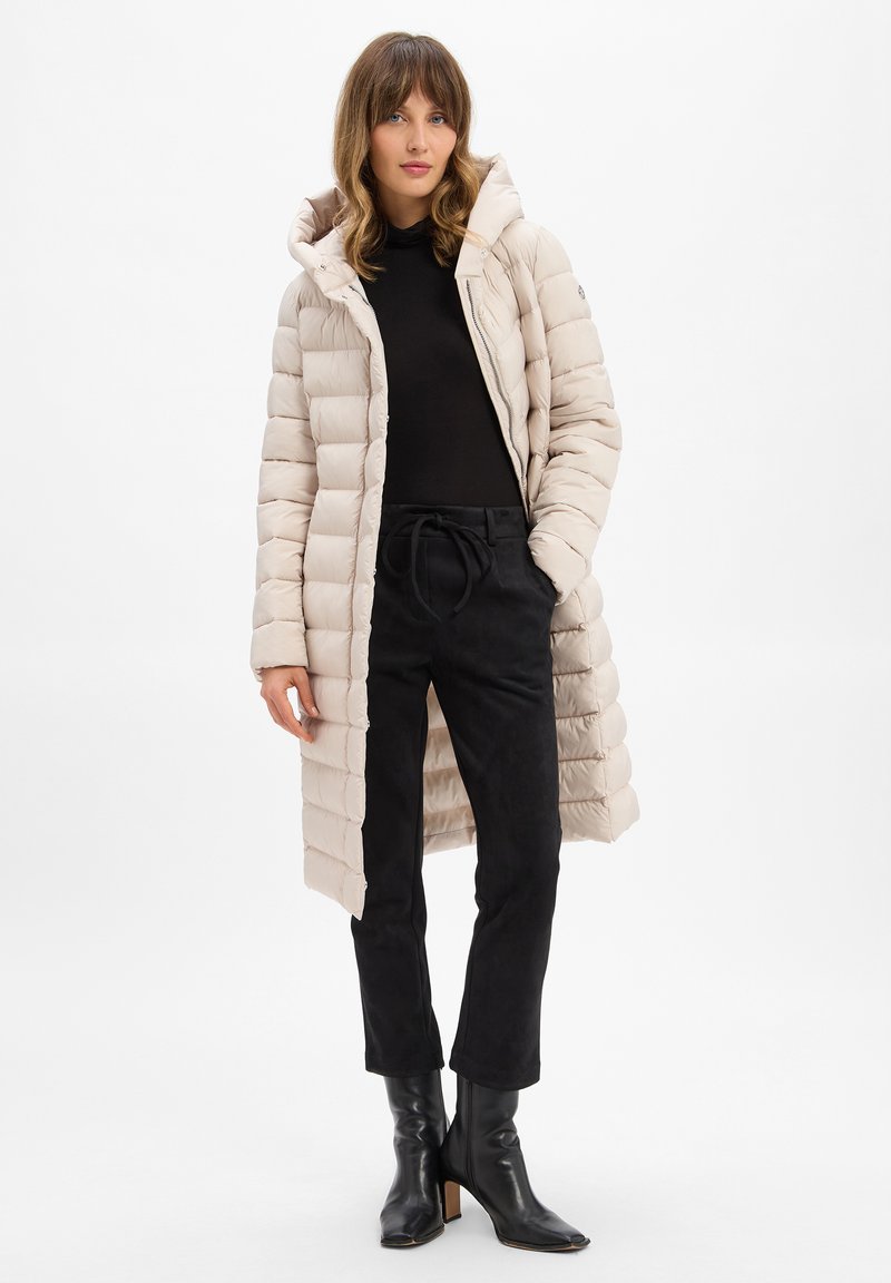 Beige Pufferjacke mit Kapuze, gestepptem Design, Frontreißverschluss und seitlichen Taschen, kombiniert mit schwarzen Wildlederhosen und schwarzen Stiefeletten mit Absatz.