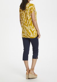 Keltainen lyhythihainen paita, jossa on valkoinen kukkakuosi, yhdistettynä tummansinisiin nilkkapitkiin housuihin ja vaaleanruskeisiin platform-sandaaleihin, joissa on nilkkaremmi.