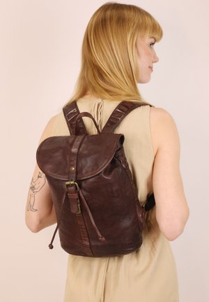 Blonde vrouw in een beige mouwloos jurkje draagt een donkerbruin leren rugzak met gespsluiting, en toont een katten tatoeage op haar linkerarm.