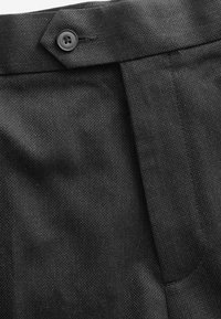 Pantalons en tissu noir avec un tissage texturé, une fermeture à boutons et une coupe ajustée. Détail notable : une section avant plissée.