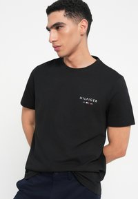 Μαύρη βαμβακερή μπλούζα με στρογγυλή λαιμόκοψη και μικρό λογότυπο "HILFIGER 1985" στην αριστερή πλευρά του στήθους με άσπρες, κόκκινες και μπλε λεπτομέρειες.